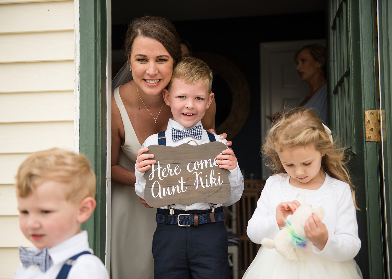 bride smiles along with her baby groomsmen. the baby groomsmen is holding a sign that reads "here comes aunt kiki"