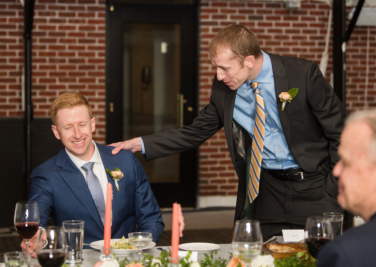 groom smiles as one of his groomsmen shares a speech during their wedding reception