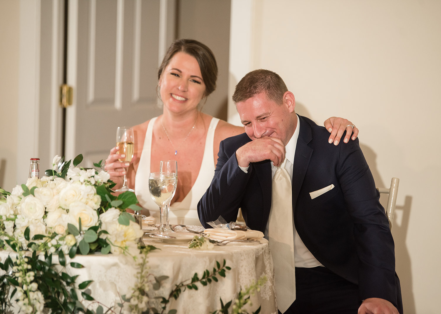 bride and groom sitting down smiling listening to the speeches from their wedding party