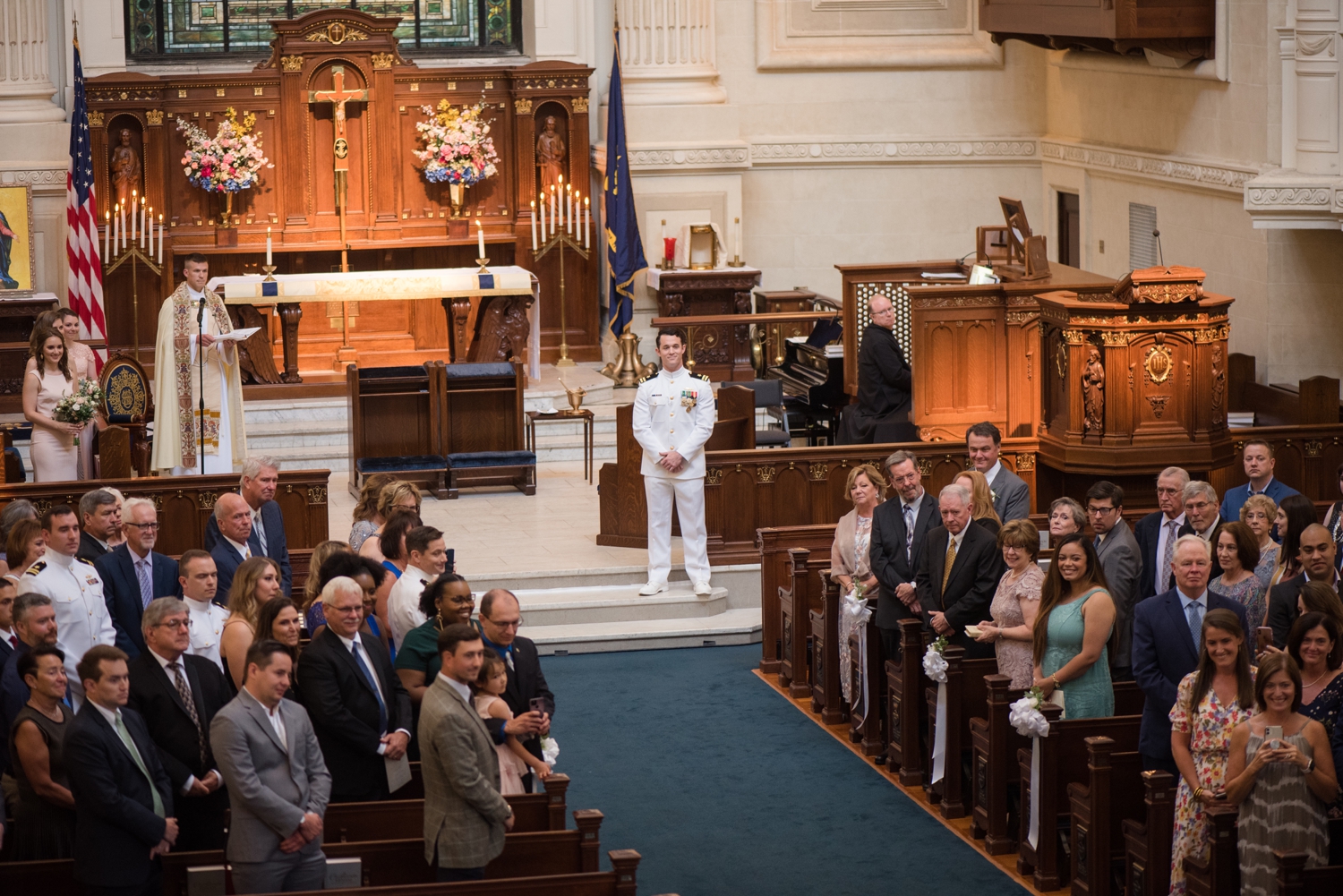 The groom waits for the bride at the end of the aisle, guests watch as she walks with her father.