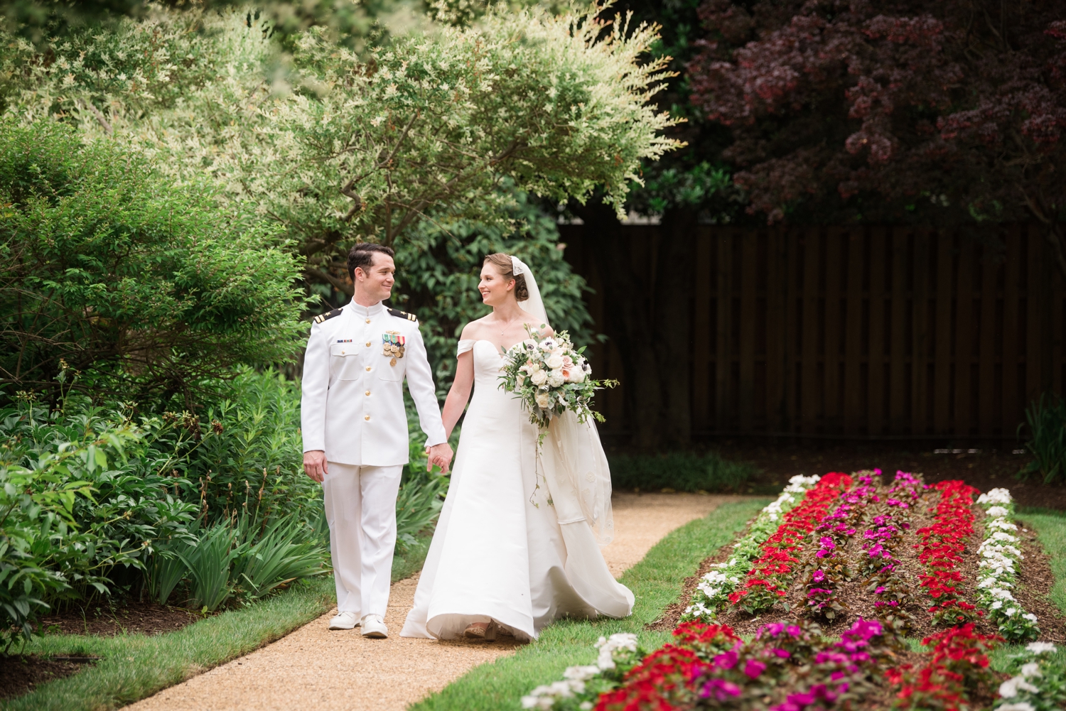 bride and groom walk hand in hand staring at each other with red flowers in the right of the frame