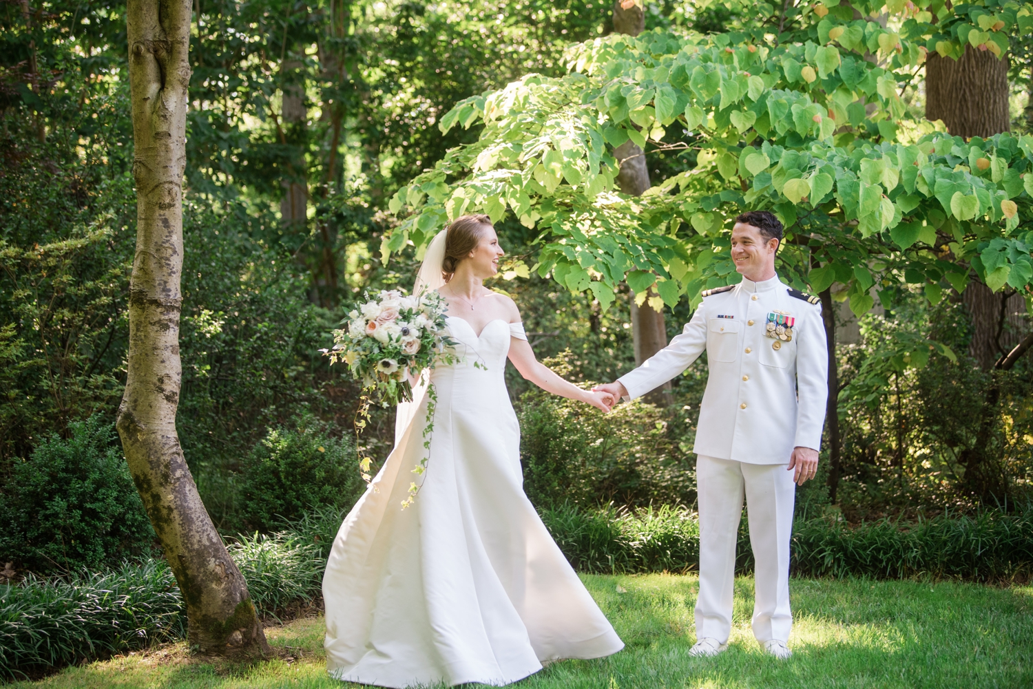 Bride and groom stand a few feet apart looking at each other and holding hands.