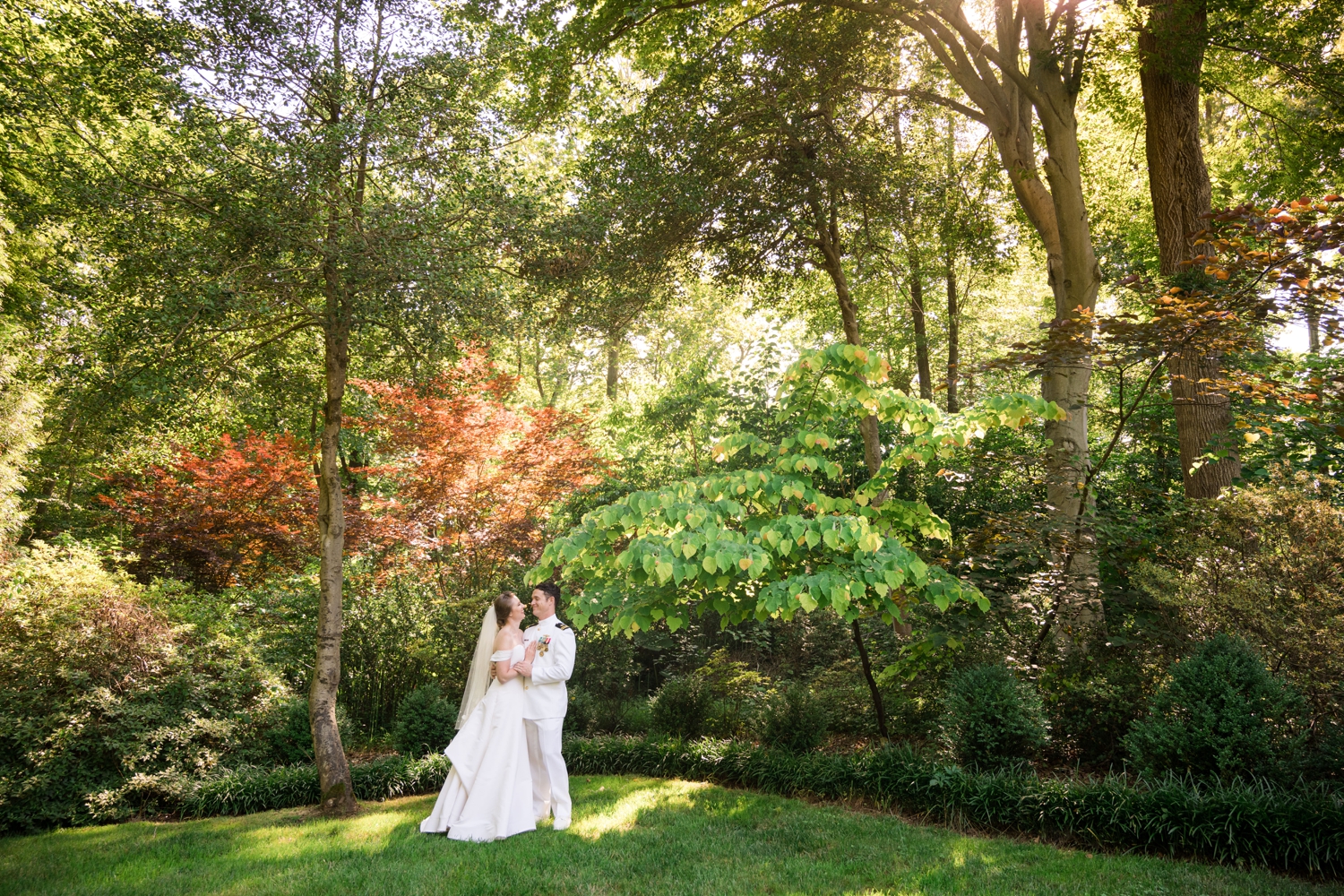 Bride and groom stand in the gardens with sunbeams lighting up the trees behind them