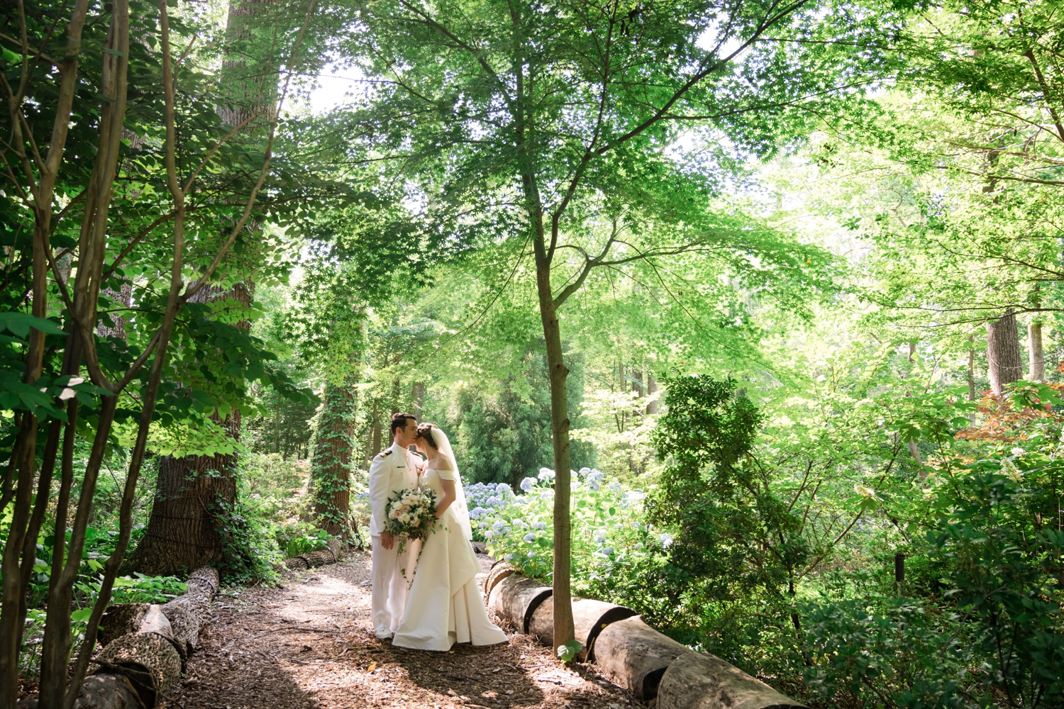 Bride and groom kiss in the garden path with the sun lighting up the trees behind them