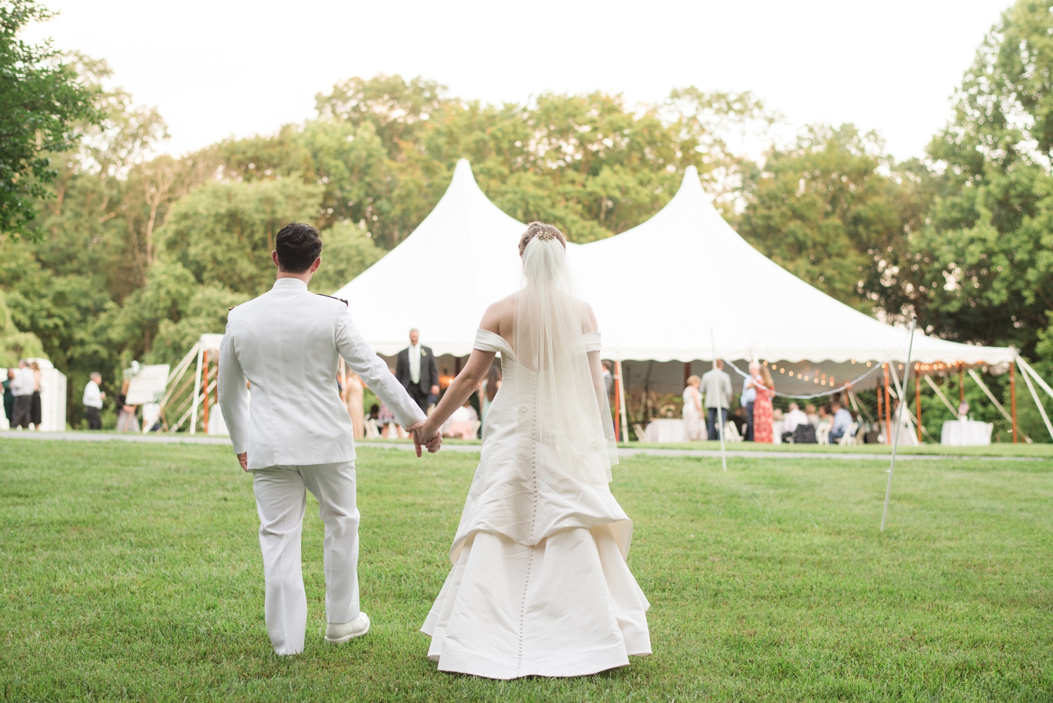 bride and groom stand with their backs to the camera facing the white tent where their lively wedding reception is taking place.