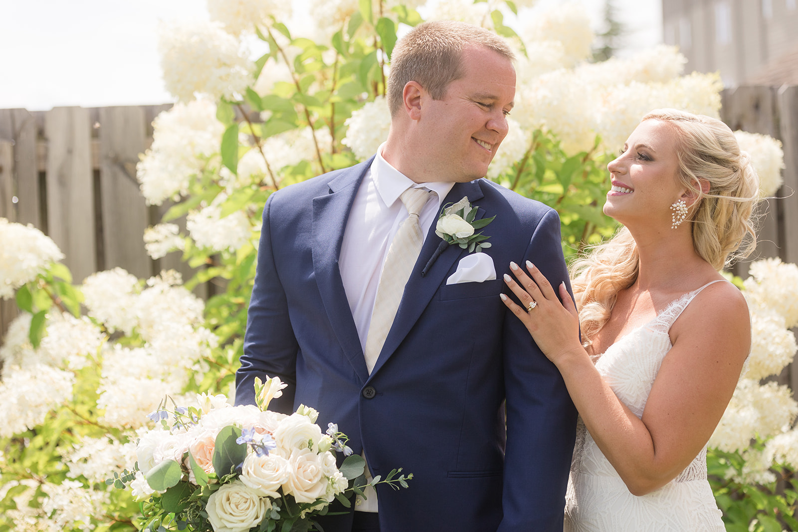 groom looking back at smiling bride in front of white flowering bushes