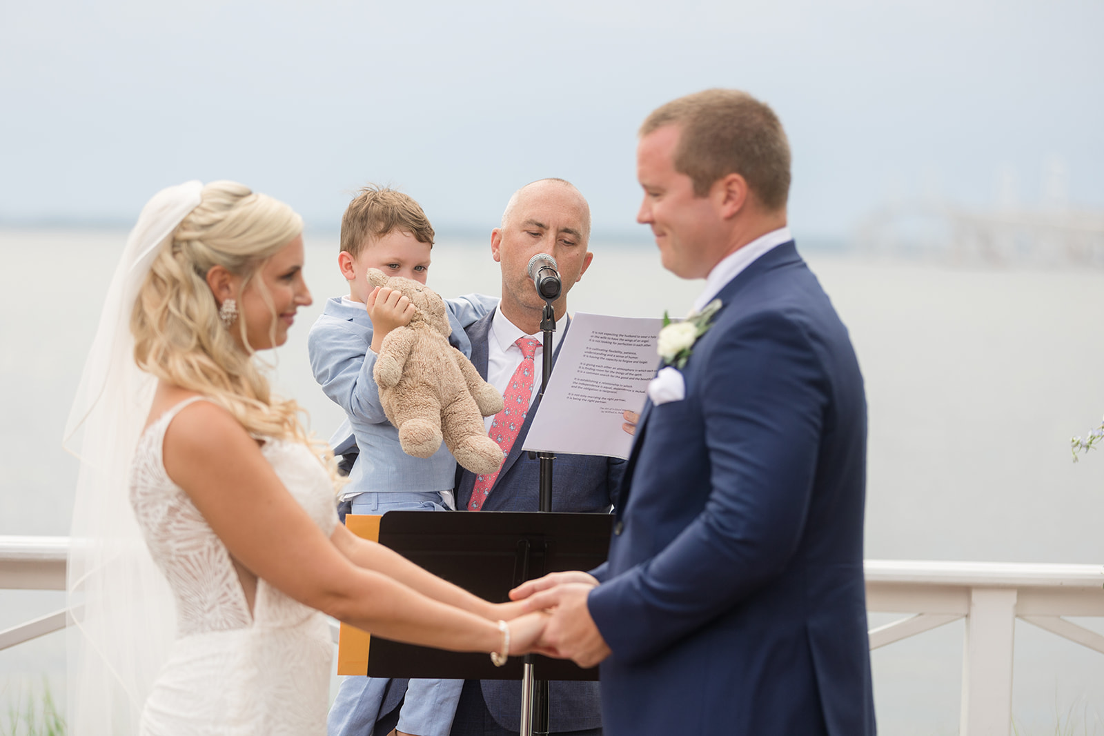 bride and groom hold hands during ceremony while officiant holds their son with his teddy bear