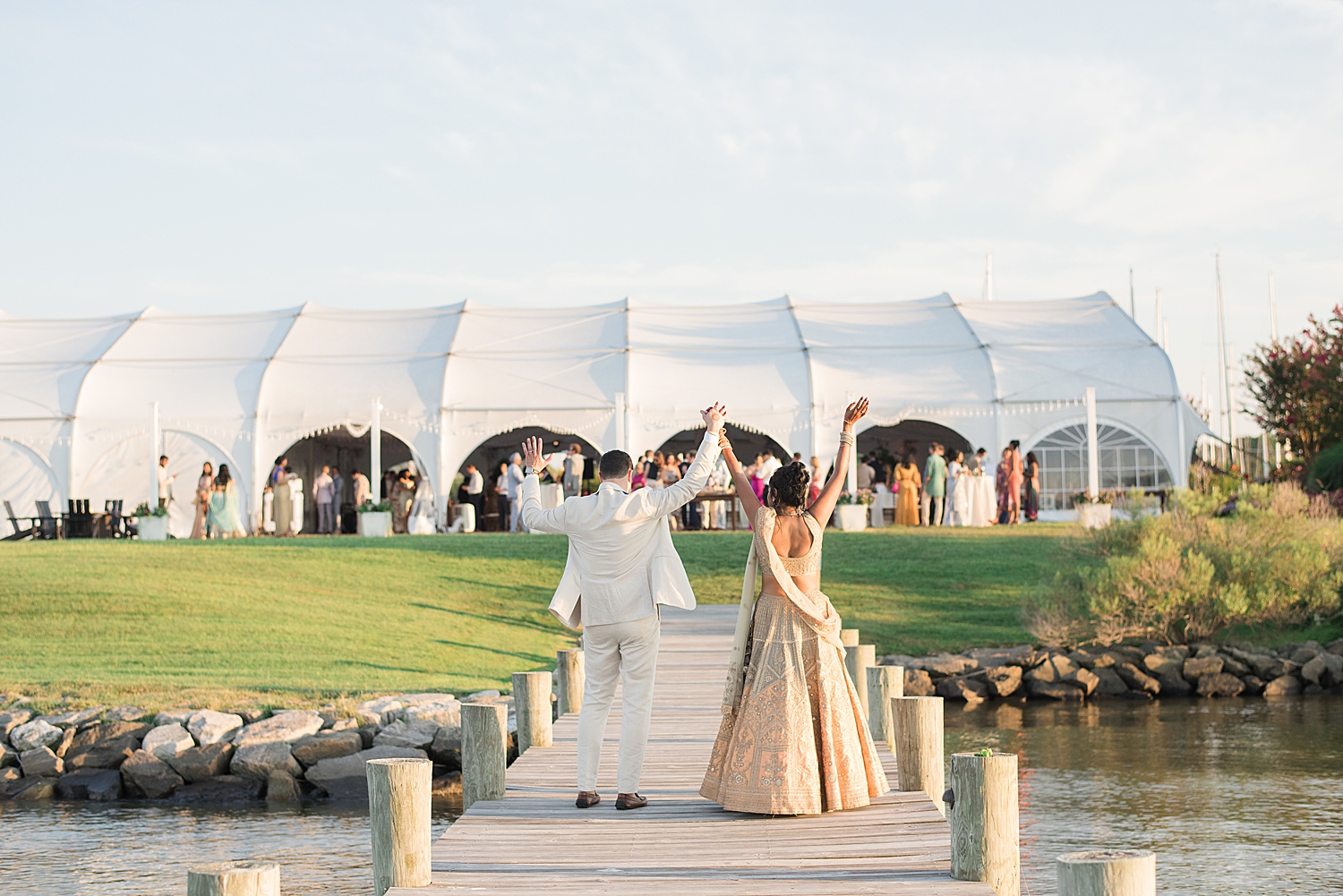 bride and groom cheer at guests under tent from pier