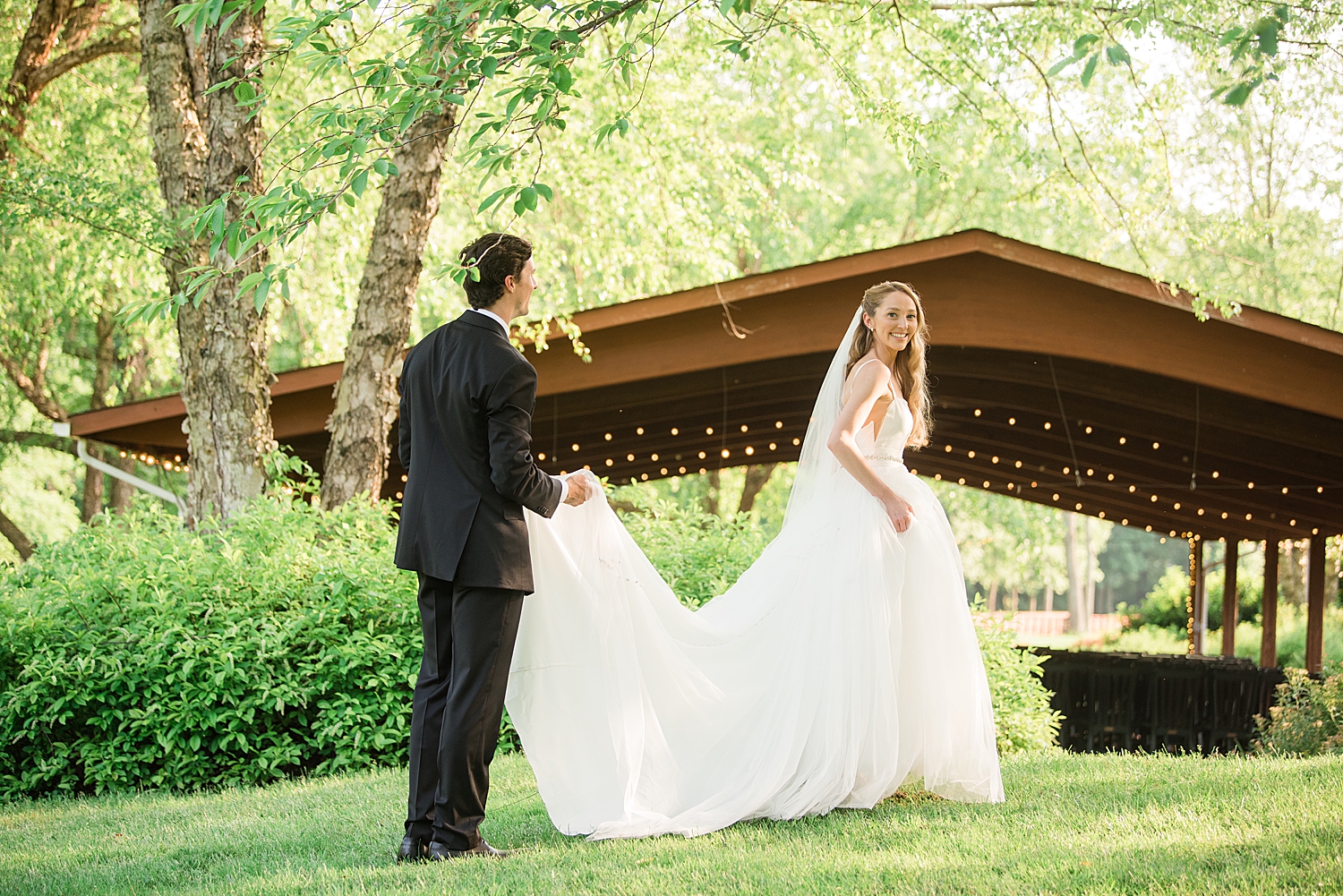 groom holds bride's train as they walk through grass