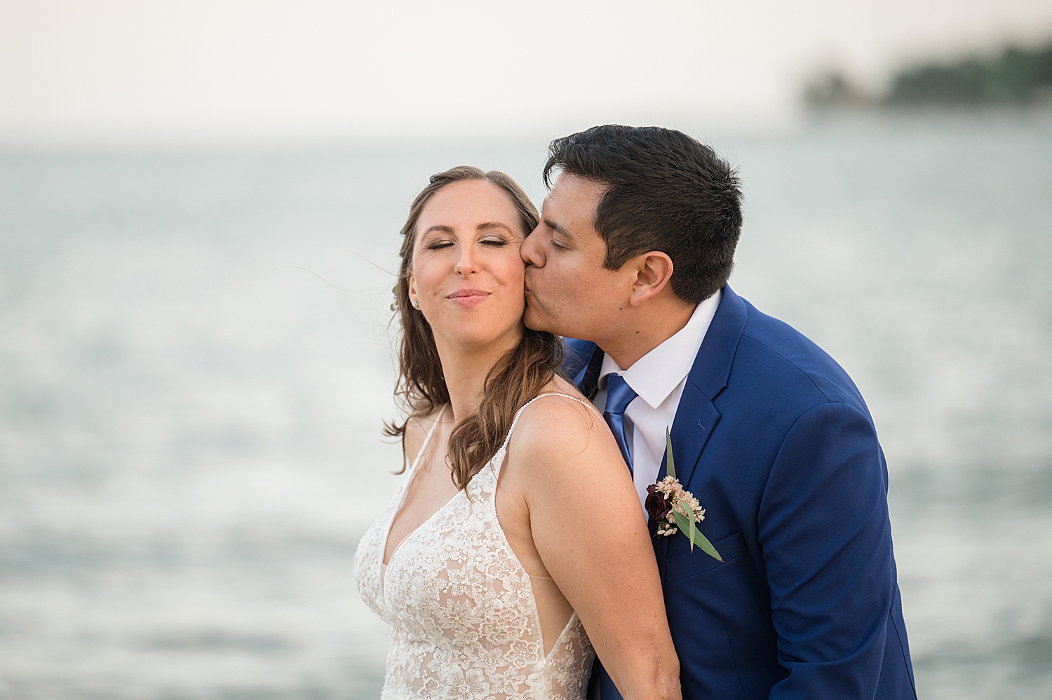 groom kissing bride's cheek with bay in the background