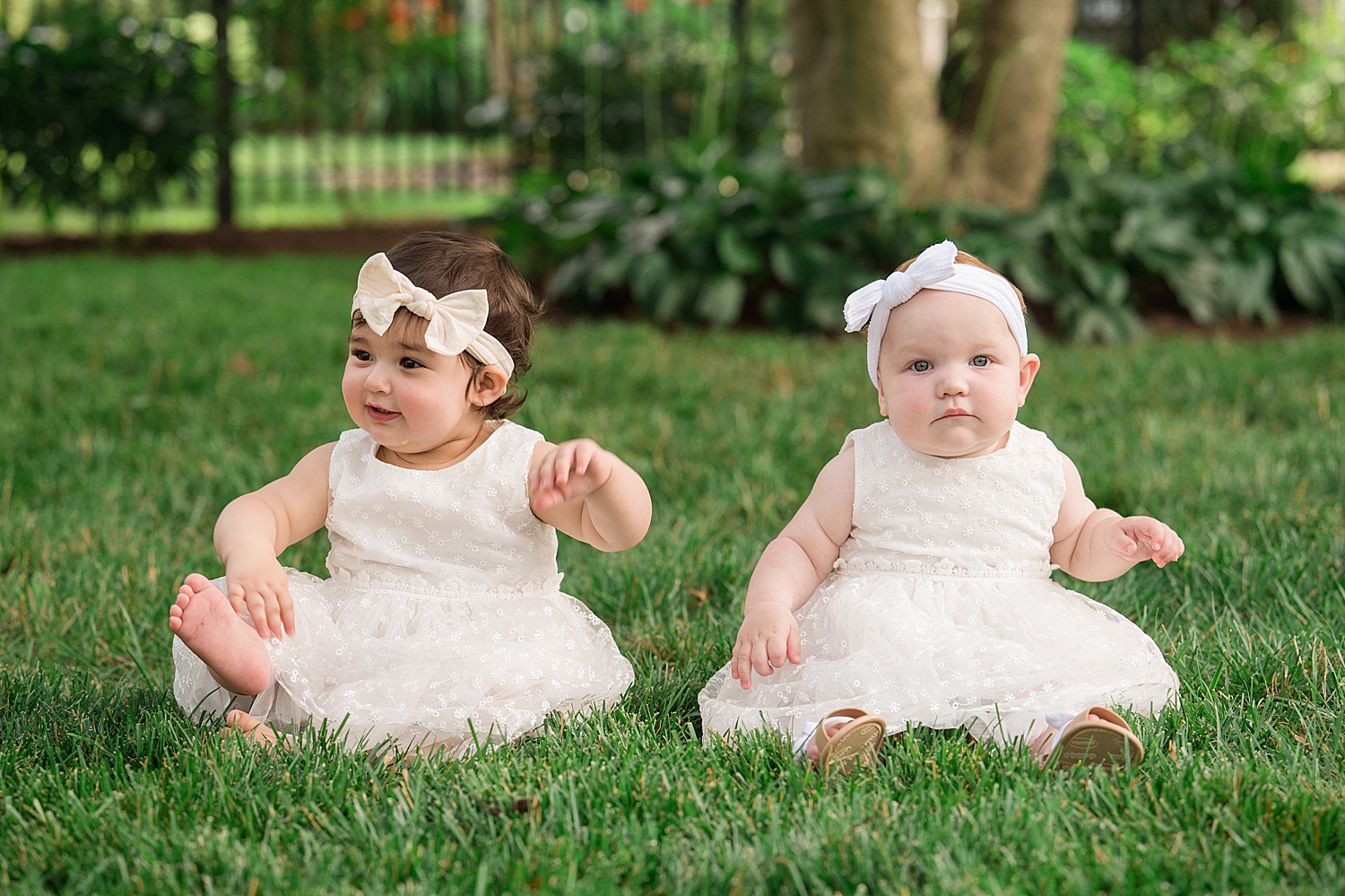 two babies dressed in white sit in the grass