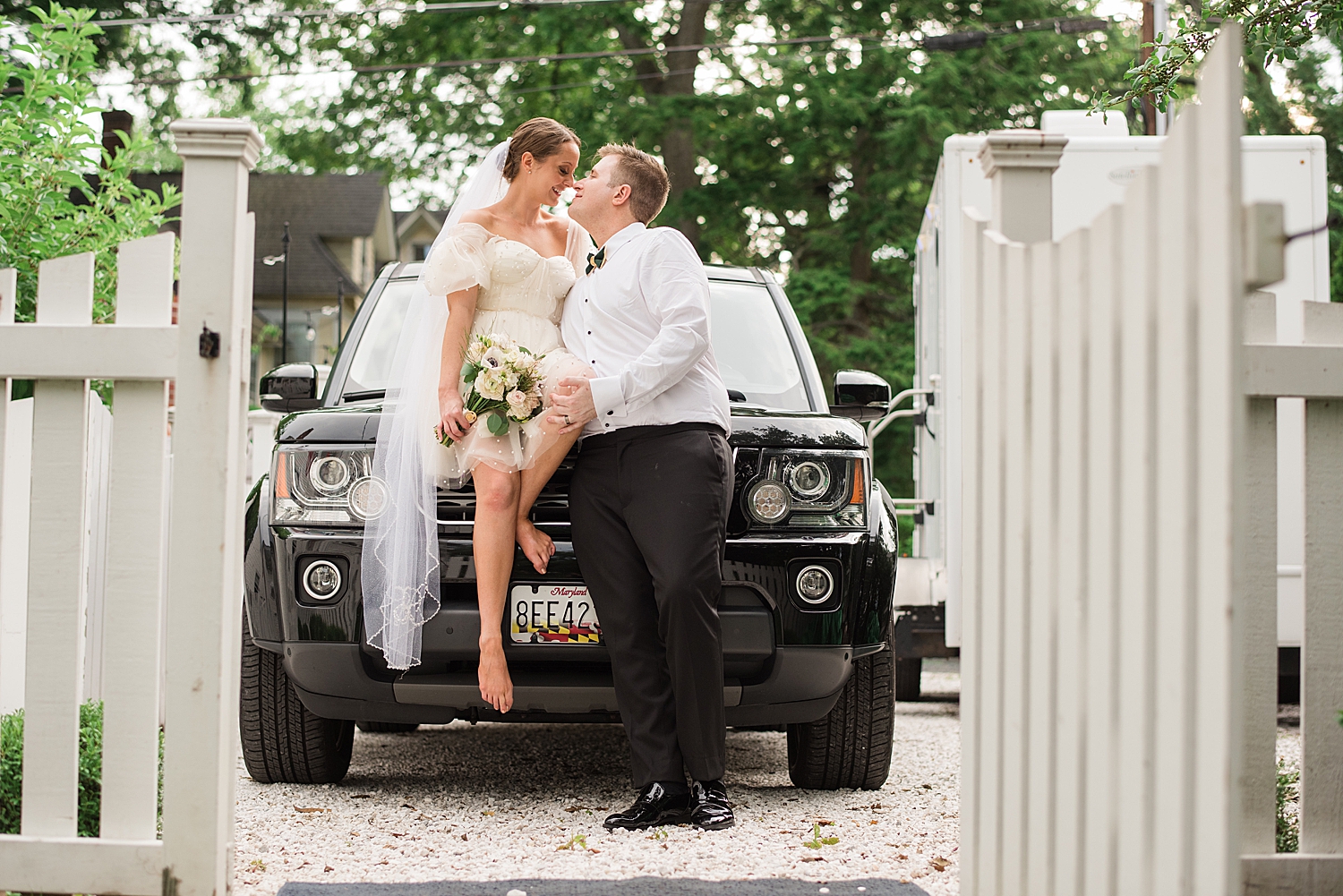 bride sits on hood of black car, kissing groom