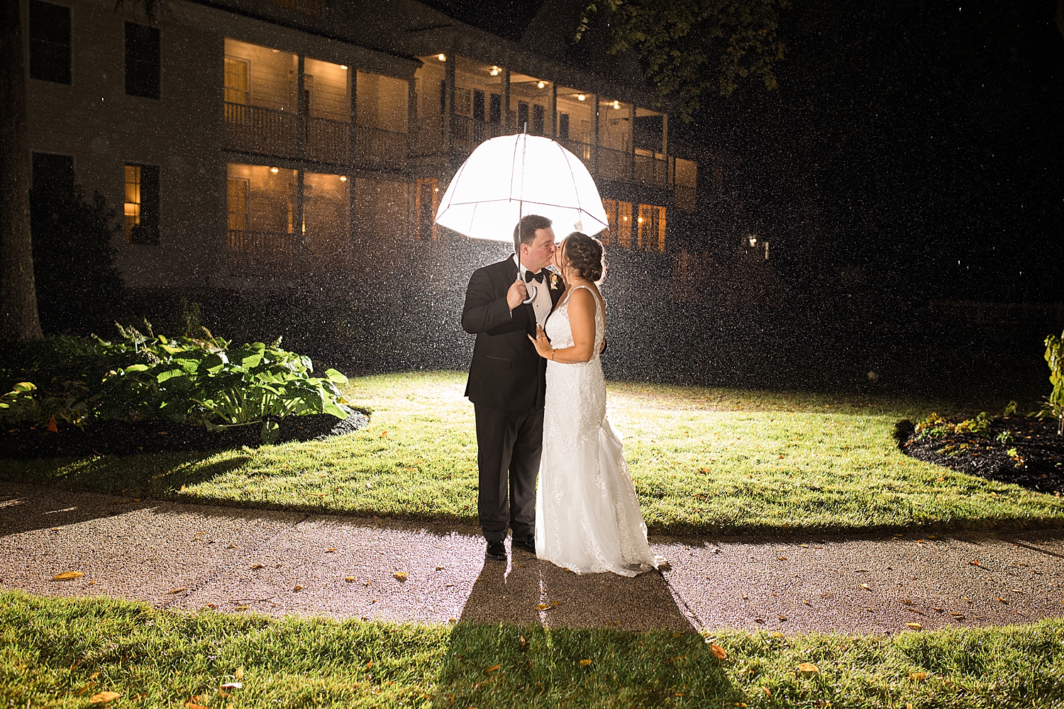 bride and groom kiss in rain with umbrella, night shot, illuminated from behind