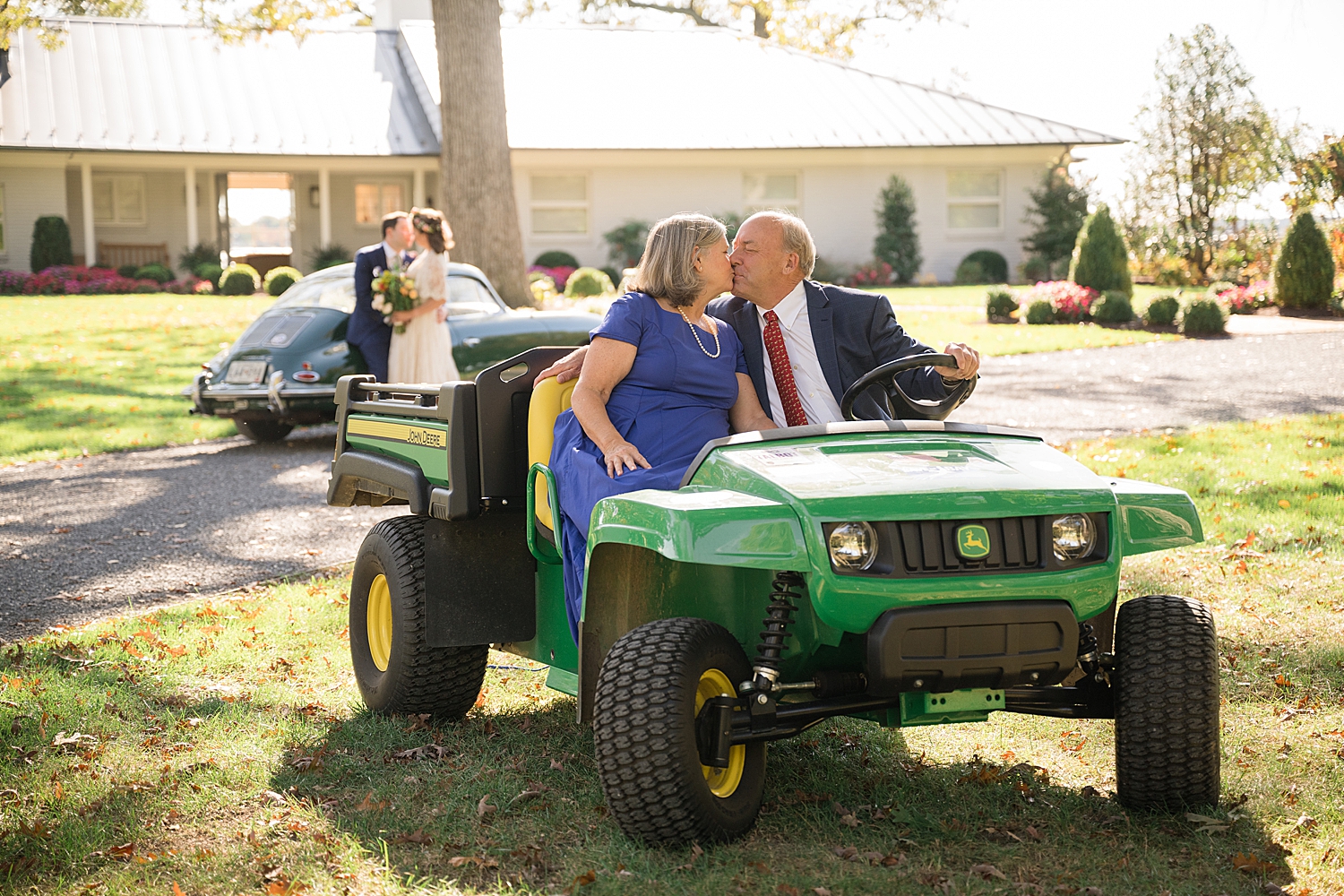 bride and groom kiss in front of green vintage car while bride's parents kiss on john deere in foreground